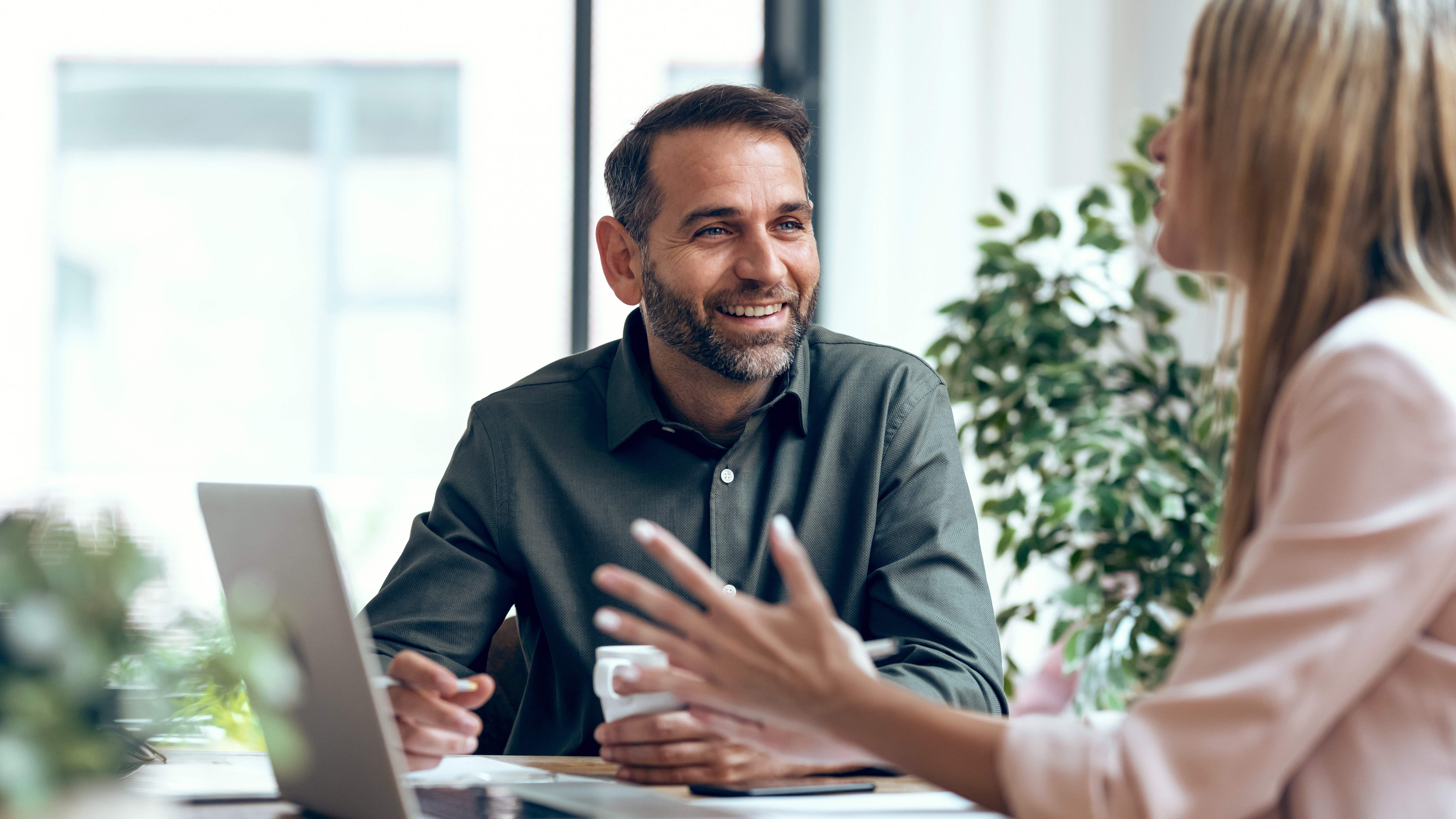 Man and woman sitting at a table in conversation, with the man visible from the front and the woman holding a laptop in front of her.