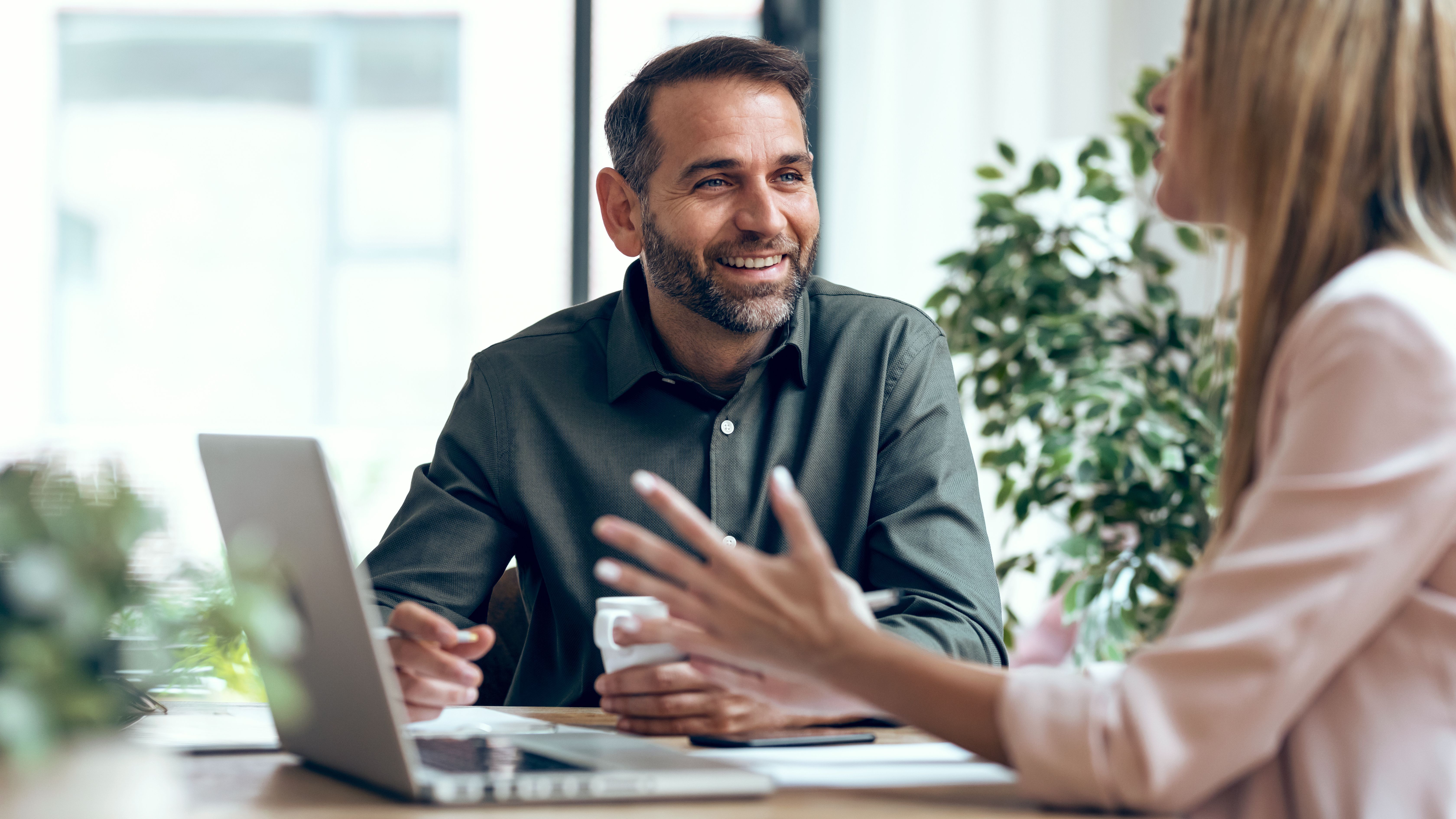 Mann und Frau an Tisch sitzend im Gespräch, wobei der Mann von vorne sichtbar ist und die Frau einen Laptop vor sich hat.
